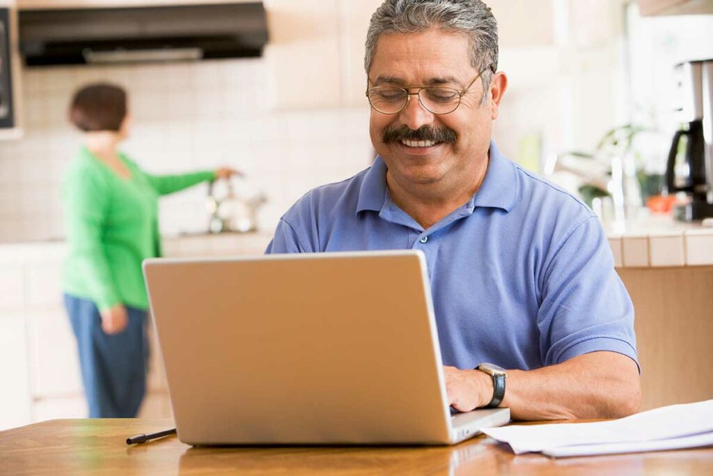 man-in-kitchen-with-laptop-smiling-with-woman-in-background-SBI-301052669